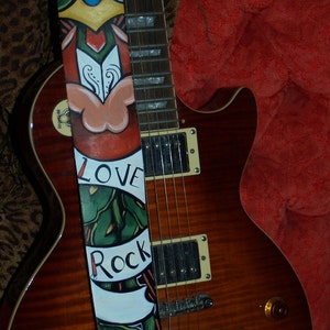 Może przedstawiać: Brązowa gitara elektryczna z malowanym paskiem. Pasek ma czarne tło z kolorowymi wzorami i napisami "Live Love Rock".