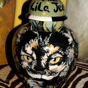 Puede incluir: Un tarro de cerámica con un diseño pintado en verde, negro y blanco. El tarro presenta un gato negro y blanco con ojos verdes y las palabras "Lila Jean" en la tapa.