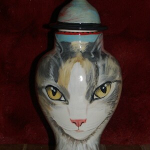 Puede incluir: Un jarrón de cerámica con forma de gato, con un diseño de gato atigrado gris, blanco y marrón. El jarrón tiene una tapa con un patrón de lunares negros y blancos.