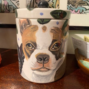 Könnte beinhalten: Weiße Keramik-Keksdose mit einem gemalten Bild eines französischen Bulldoggen. Der Hund hat braunes, weißes und schwarzes Fell. Die Dose hat einen schwarzen Knopf auf dem Deckel und grüne Blätter und blaue Punkte, die auf den Deckel gemalt sind.