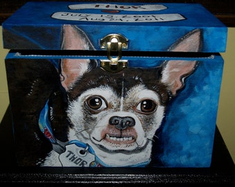 May include: A blue wooden box with a brass latch, painted with a portrait of a chihuahua dog. The dog has brown and white fur and is wearing a blue collar with a tag that says "Thor". The box has a white label on the top that says "Thor July 15, 2001 Aug 24, 2011".