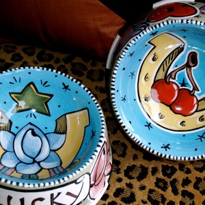 May include: Two ceramic pet bowls with blue backgrounds and white rims. One bowl has a horseshoe with cherries and the other has a horseshoe with a blue flower and a star. Both bowls have the word "LUCKY" written on the front.