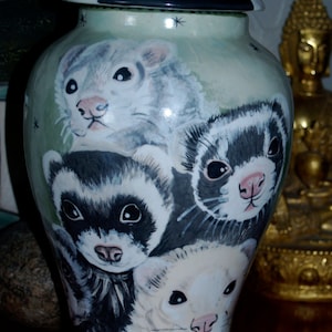 May include: A ceramic urn with a green background and a black lid. The urn is decorated with a painted image of four ferrets. The lid has the words "Grant Ferre" written on it.