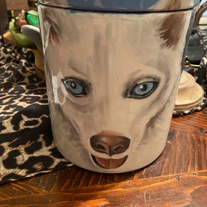 Pode incluir: Jarro de biscoitos de cerâmica branca com uma imagem pintada de um rosto de cão branco. O cão tem olhos azuis e uma língua rosa. O jarro tem uma tampa azul com um desenho floral branco.