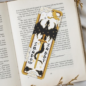 Puede incluir: Marcapáginas bordado con una espada y la palabra "SHADY" en fuente gótica. El marcapáginas presenta una silueta de castillo negro, detalles dorados y un diseño de dragón. Está colocado sobre un libro abierto.