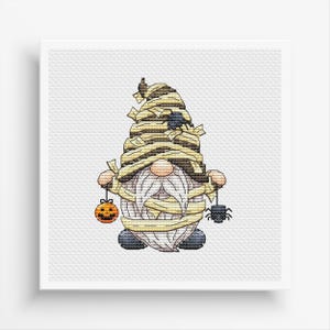 Op de afbeelding: Een witte, grijze en bruine gnome is gewikkeld in verband en houdt een kleine oranje pompoen en een zwarte spin vast. De gnome draagt een zwarte hoed.