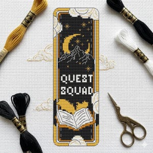Może przedstawiać: Czarna i złota zakładka do krzyżyków z napisem "QUEST SQUAD". Zakładka przedstawia księżyc w kształcie półksiężyca, góry, gwiazdy i wzór otwartej książki. Wokół zakładki znajdują się nici do haftu i nożyczki.