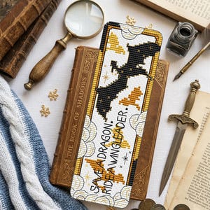 May include: A rectangular cross-stitch bookmark featuring a black dragon silhouette and gold dragon shapes against a white background with cloud outlines. The text reads "SAVE A DRAGON: RIDE A WNGLEADER." with star accents. The design is bordered by a gold frame.