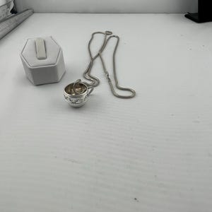 Puede incluir: Un collar de cadena de plata con un colgante de taza de plata. La taza tiene un diseño detallado.