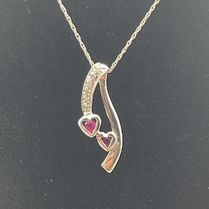 Puede incluir: Collar con colgante de plata con un diseño curvo. El colgante presenta dos piedras preciosas rojas en forma de corazón y una sección incrustada con pequeñas piedras transparentes. El collar cuelga de una delicada cadena de plata.