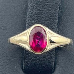 May include: A gold ring featuring an oval, deep red gemstone. The ring's band has a simple, elegant design, with a smooth, polished finish. The gemstone is the focal point, set in a classic bezel setting.