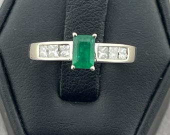 May include: A silver ring featuring a rectangular emerald center stone, flanked by square-cut diamonds. The ring is set on a black display surface, showcasing the jewelry's design and sparkle.