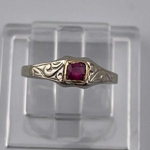 Peut inclure: Une bague argentée ornée d'une pierre précieuse rouge foncé de taille carrée, sertie dans une monture dorée. L'anneau présente un motif de volutes en relief de chaque côté de la pierre centrale, offrant un aspect texturé.