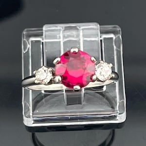 May include: A silver-toned ring featuring a large, round, vibrant red center stone held by six prongs, flanked by two smaller, round, clear white stones on either side. The band is smooth and metallic, showcasing a classic three-stone design.