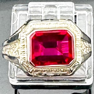 May include: A ring featuring a large, rectangular, faceted vibrant red gemstone set in a gold-toned metal band. The metal setting surrounding the stone displays intricate, raised, textured detailing along the edges and the sides of the band.
