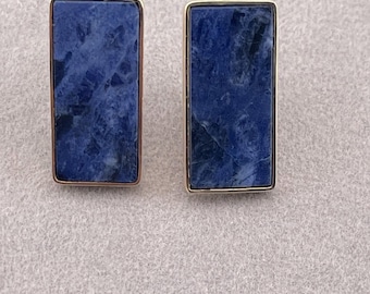 Può includere: Un paio di orecchini a bottone rettangolari. Ogni orecchino presenta una pietra blu scuro con venature bianche, incastonata in una montatura in metallo color oro. Gli orecchini sono esposti su una superficie grigio chiaro.