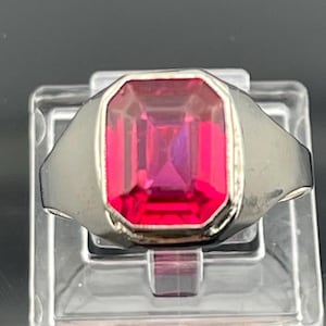 以下が含まれることがあります： 銀色の金属製リング。中央にはエメラルドカットが施された大きな長方形の鮮やかなピンク色の宝石が、滑らかで光沢のあるベゼルセッティングで固定されています。リングのバンド部分は幅が広く、後ろに向かってわずかに細くなっており、すっきりとした金属の質感が特徴です。
