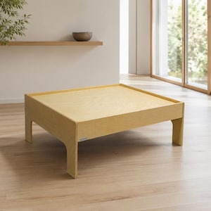 May include: A square, low-profile coffee table constructed from light-toned plywood. The table features a flat, smooth surface with a raised edge border and four sturdy, integrated legs with arched cutouts at the base, showcasing a natural wood grain.