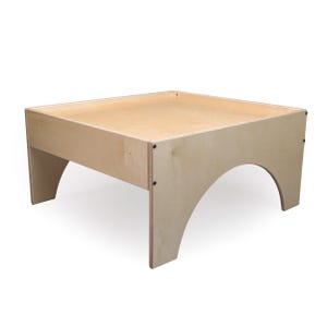 May include: A square light-colored wooden sensory table with a flat recessed surface and raised edges. The table features four sturdy legs, each designed with a distinct arched cutout on the side, and is constructed with visible metal screws.