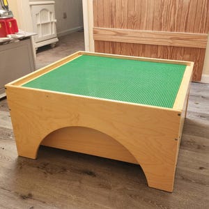May include: A wooden play table with a green surface designed for building blocks. The table has a light wood finish and an arched design on the sides. The table is rectangular and sits on a wooden floor.