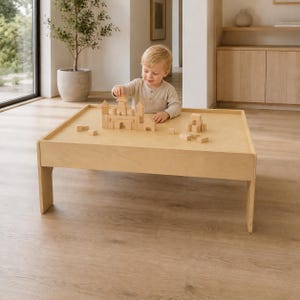 May include: A light-colored wooden play table with a raised edge, featuring a set of natural wood building blocks arranged into a castle structure on the surface. The table has a smooth, flat top and sturdy legs, providing a dedicated space for play.