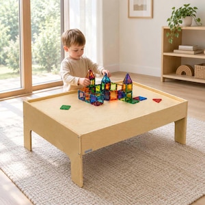May include: A light-colored wooden play table with a low profile and four sturdy legs. On the surface, a collection of translucent, multi-colored magnetic building tiles are arranged into various geometric shapes and small tower structures.