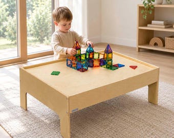 May include: A light-colored wooden play table with a low profile and four sturdy legs. On the surface, a collection of translucent, multi-colored magnetic building tiles are arranged into various geometric shapes and small tower structures.