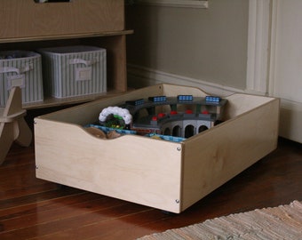 May include: A light-colored wooden toy box on wheels, filled with a toy train set. The box has a curved cutout on one side for easy access. Other wooden shelves and storage bins are visible in the background.