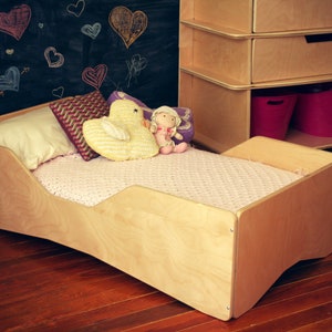 May include: A light wood toddler bed with a pink blanket and two stuffed toys. The bed has a high back and sides to prevent the child from falling out.