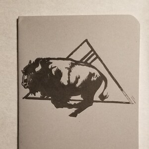May include: A tan notebook with a black illustration of a bison running through a triangle. The bison is detailed with visible fur and horns. The triangle has three vertical lines inside. The notebook has rounded corners.