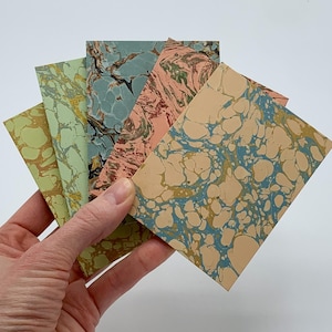 May include: A hand holds a fanned stack of rectangular cards with marbled paper designs. The cards feature a variety of colors, including green, blue, pink, and tan, with swirling patterns. The cards are likely for stationery or art projects.