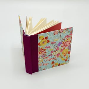 May include: A small, floral-patterned journal with a burgundy spine and red endpapers. The cover features a blue background with pink, yellow, and red flowers.
