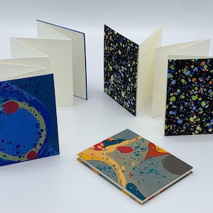 May include: Four small, folded paper notebooks with marbled covers. The covers feature different color combinations of blue, green, yellow, red, and black.