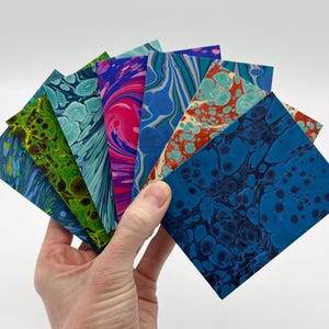 May include: A hand holding a fan of colorful, marbled paper squares. The squares display a variety of patterns in shades of blue, green, pink, and orange. The marbled effect creates a unique, artistic look.