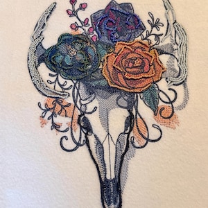 May include: An embroidery design featuring a central animal skull outlined in dark thread, adorned with a cluster of flowers in shades of orange, blue, and purple. The skull is flanked by two curved horns embellished with silver-toned beads.