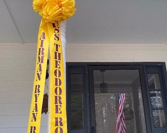 May include: A yellow ribbon with the text "USS THEODORE ROOSEVELT AIRMAN RYAN RIKE" in maroon lettering hangs from a white porch post. A small dog is visible through a glass door behind the ribbon.