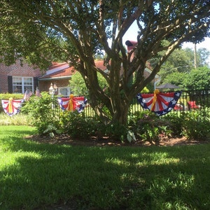 May include: A brick house with a large tree in the front yard. The tree is decorated with red, white, and blue bunting with yellow ribbons. There is a black metal fence in the background.