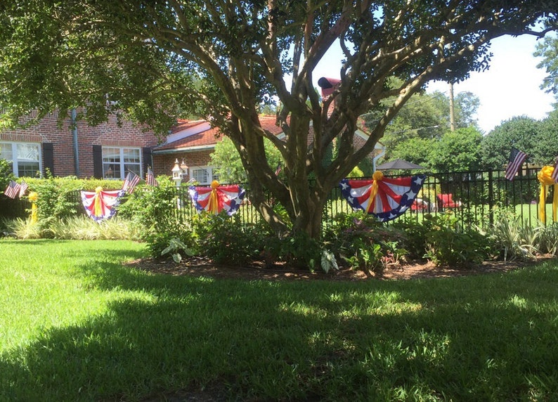 May include: A brick house with a large tree in the front yard. The tree is decorated with red, white, and blue bunting with yellow ribbons. There is a black metal fence in the background.