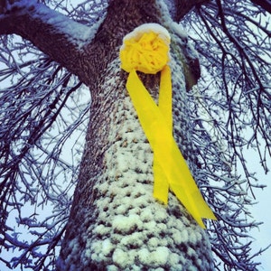 May include: A yellow ribbon tied around a tree trunk covered in snow. The ribbon is tied in a bow with a large yellow flower in the center.