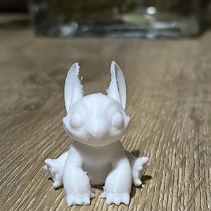 May include: White 3D printed figurine of a cartoon character with large ears and a wide smile. The character is sitting with its legs crossed and its arms resting on its knees.