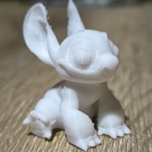 May include: A white 3D printed figurine of Stitch from the Disney film Lilo & Stitch. The figurine is sitting with its ears up and its mouth slightly open.