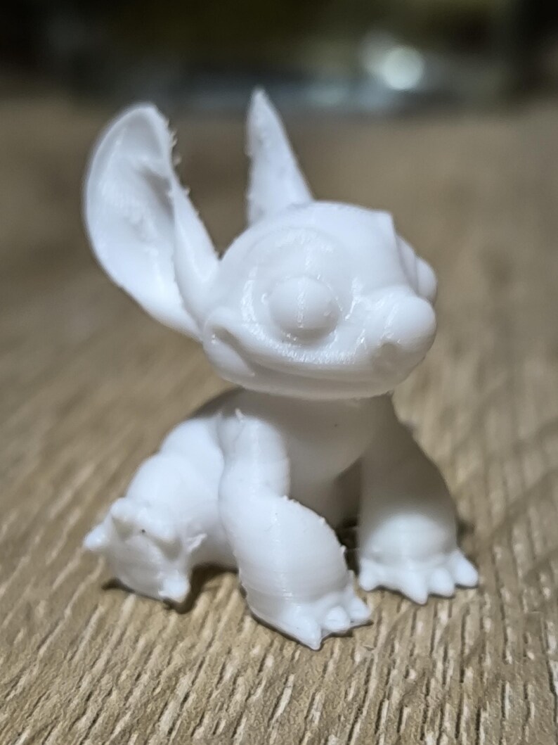 May include: A white 3D printed figurine of Stitch from the Disney film Lilo & Stitch. The figurine is sitting with its ears up and its mouth slightly open.
