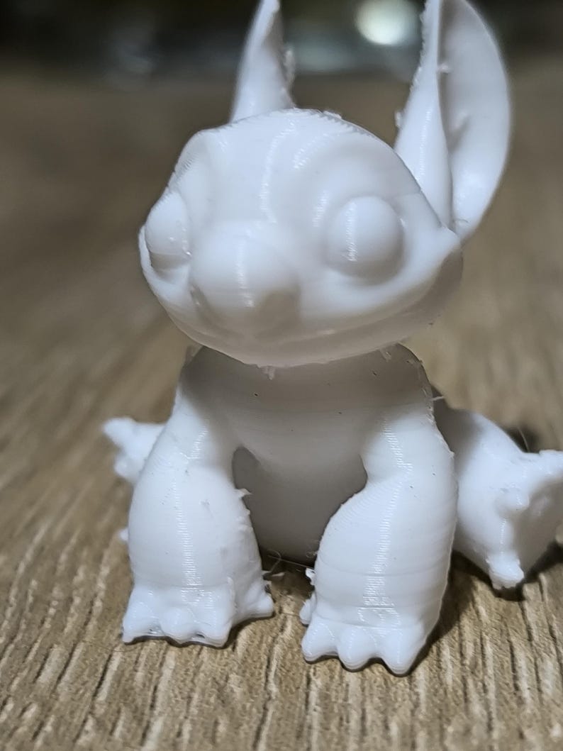 May include: White 3D printed figurine of a cartoon character with large ears, sitting with its legs crossed. The character has a wide smile and large eyes.