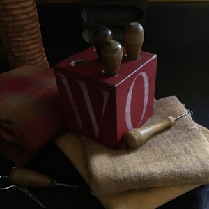 May include: A red wooden block with the letter "W" and "O" painted on it. The block has four holes in the top, each holding a wooden dowel. The block is sitting on a stack of beige fabric.
