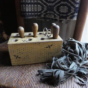 May include: A yellow wooden box with eight holes for holding tools. The box has a textured surface and is decorated with black dragonfly designs. There are four wooden tools in the box, and a pair of silver scissors. A pile of dark green leather cord is in the foreground.