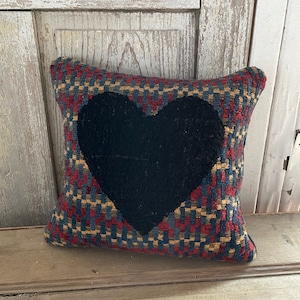 May include: Square decorative pillow with a woven pattern in red, blue, and tan. A large black heart is centered on the front. The pillow is placed on a wooden surface.