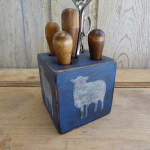 May include: A blue wooden tool holder with a white sheep design. The holder has four slots for tools, and there are three wooden tools and a pair of scissors in the slots.