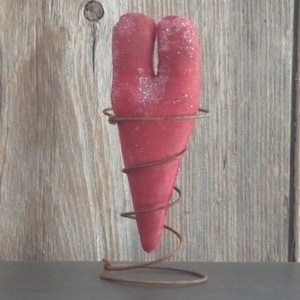 May include: A red heart-shaped decoration with a textured surface, held by a rusty metal wire stand. The heart has a split down the middle. The background is a wooden wall.