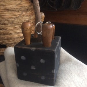 May include: A black wooden cube with white polka dots holds several wooden tools, including a pencil and wooden-handled tools. The cube sits on a white, textured surface, with a bundle of twine in the background.