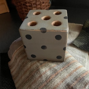 May include: A gray wooden dice-shaped holder with black dots and six holes on top. The holder is sitting on a patterned fabric with brown and tan stripes. A woven basket is in the background.
